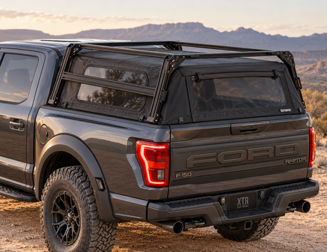 A black Ford truck with a bed rack over a soft topper cover with s side window screens installed on a dark gray Ford F-150 Raptor, parked outdoors in a desert mountain setting.